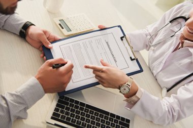 cropped view of client signing insurance claim form while doctor pointing at it in clinic with laptop and calculator clipart