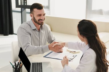 smiling client shaking hands with doctor in office with laptop and insurance claim form