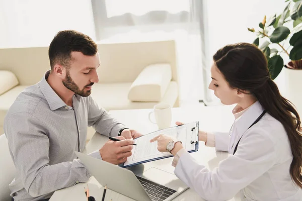 Mann Unterschreibt Versicherungsantragsformular Während Arzt Büro Mit Laptop Darauf Zeigt — Stockfoto