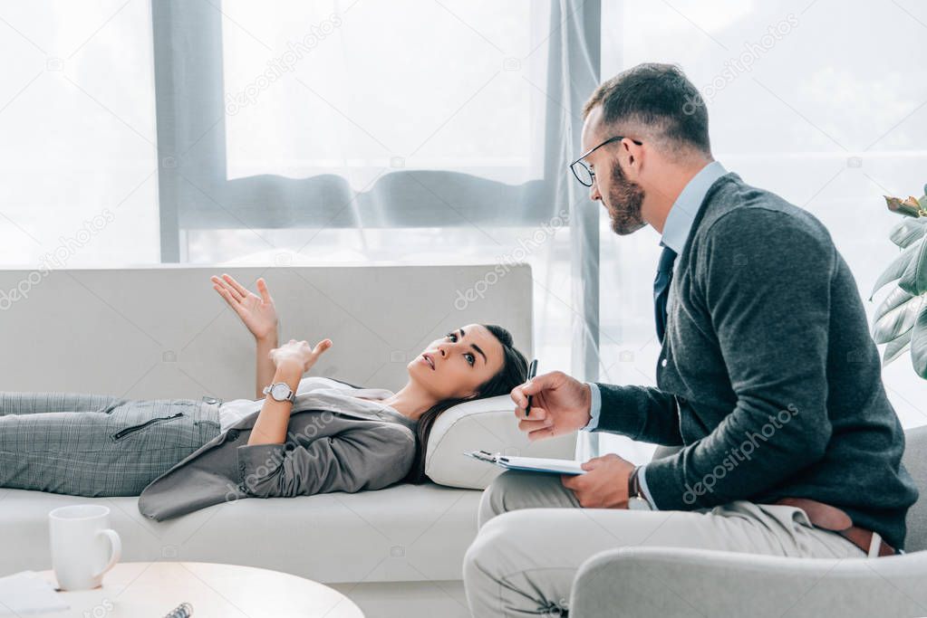 side view of patient lying on sofa and talking with psychologist in office