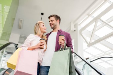 Alışveriş merkezinde yürüyen merdiven üzerinde alışveriş kağıt poşet ve kahve fincanı ile genç çiftin düşük açılı görünüş 