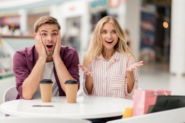 surprised young woman gesturing by hands while her shocked boyfriend sitting near at table with coffee cups in cafe clipart