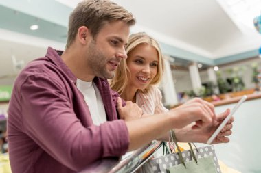 Alışveriş merkezinde dijital tablet kullanarak kağıt torbalar ile genç çift 