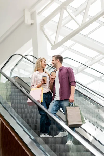 在购物中心的自动扶梯上用纸袋和咖啡杯对购物者微笑的情侣的低角度看 — 图库照片