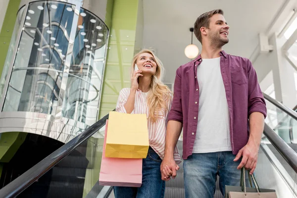 低角度的妇女与购物袋谈论智能手机 而她的男朋友站在附近的自动扶梯在购物中心 — 图库照片