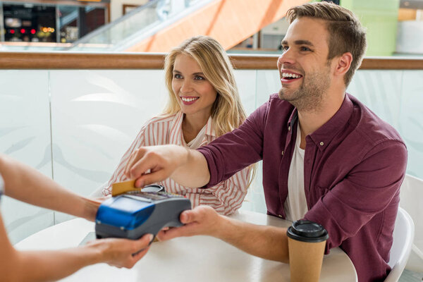 smiling young man paying by credit card on terminal while his girlfriend sitting near at table with coffee cups in cafe 