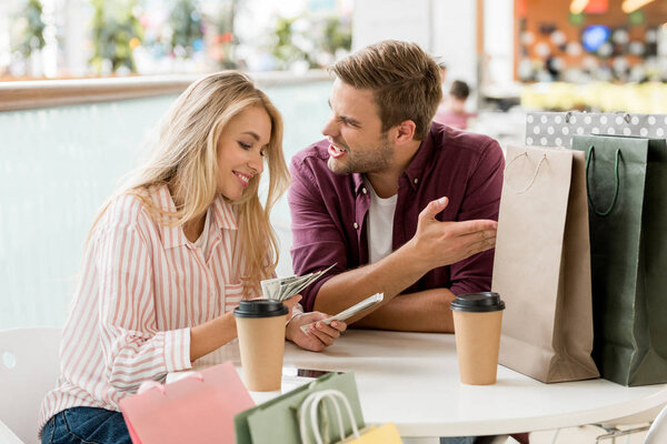 smiling young woman counting cash money after shopping while her upset boyfriend sitting near at table with coffee cups and paper bags in cae 