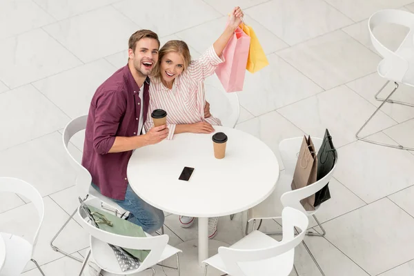 高角度的夫妇显示购物袋坐在桌子上 用一次性杯子和智能手机与空白屏幕在咖啡馆 — 免费的图库照片