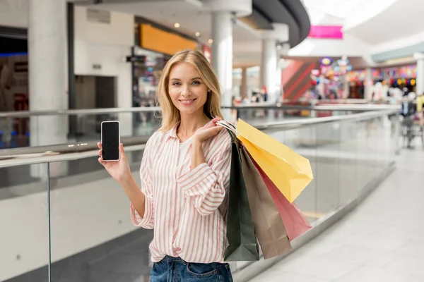 ショッピング モールで空白の画面を持つスマート フォンを示すペーパー バッグで幸せな若い女 — ストック写真