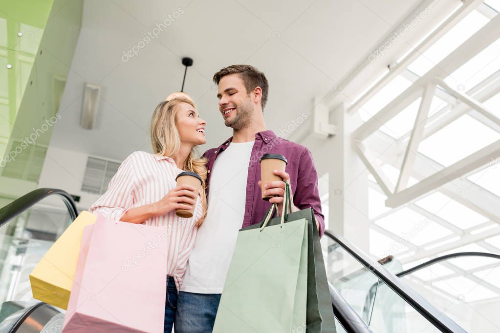 low angle view of young couple of shoppers with paper bags and coffee cups on escalator at mall 