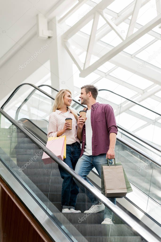 low angle view of smiling couple of shoppers with paper bags and coffee cups on escalator at mall 
