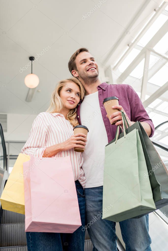 low angle view of couple with disposable coffee cups and paper bags on escalator at mall 