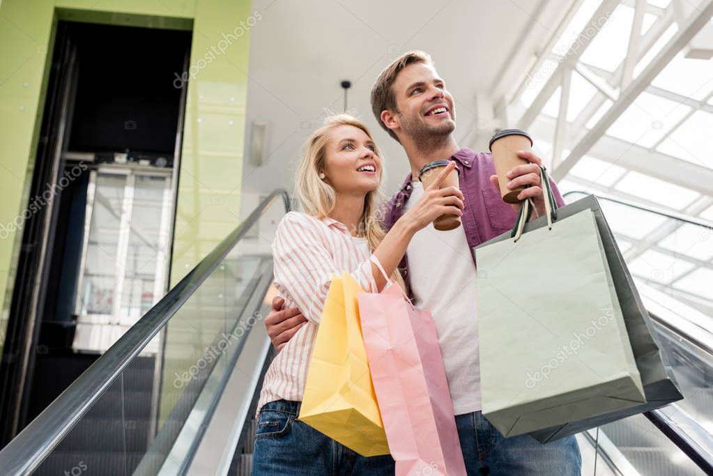 young woman with paper cup of coffee and shopping bags pointing to boyfriend on escalator at mall 