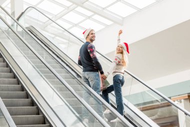 young woman in christmas hat with raised arms looking at camera while her boyfriend standing near on escalator  clipart