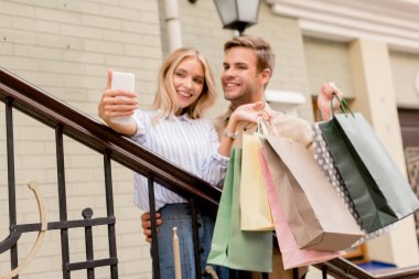 Genç çift selfie smartphone mağaza City yakınındaki sokak alarak kağıt torbalar ile gülümseyen 