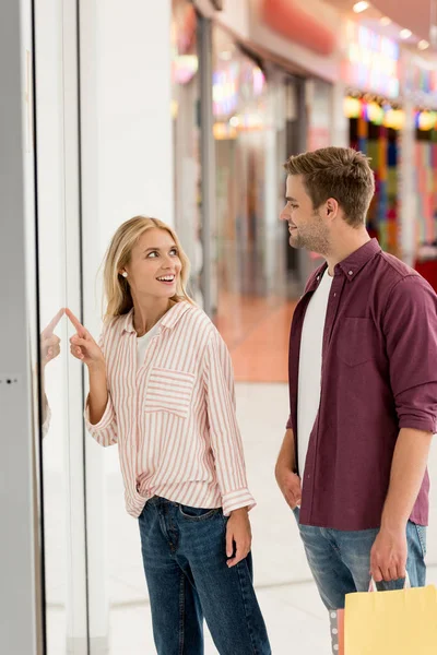 微笑的女人指着手指在橱窗的男朋友与购物袋在购物中心 — 免费的图库照片