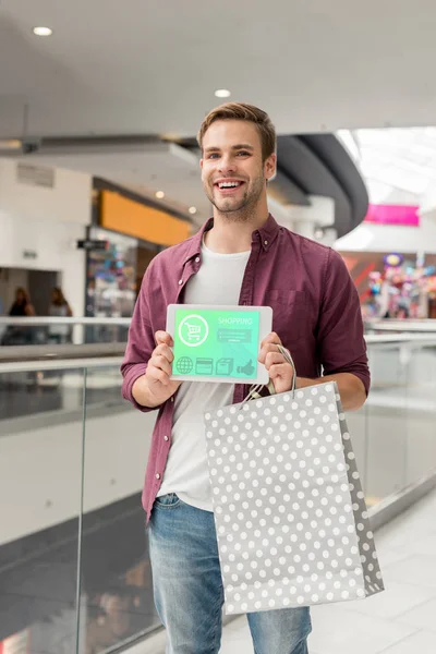 smiling man with paper bag showing digital tablet with shopping on screen at mall