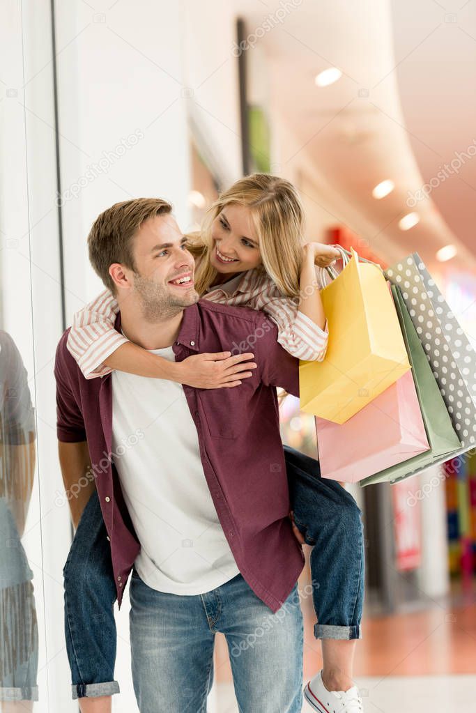 smiling man giving piggyback ride to girlfriend with shopping bags at mall 