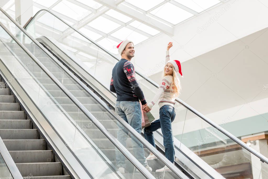 young woman in christmas hat with raised arms looking at camera while her boyfriend standing near on escalator 