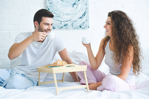 happy young couple in pajamas drinking coffee and smiling each other while sitting on bed