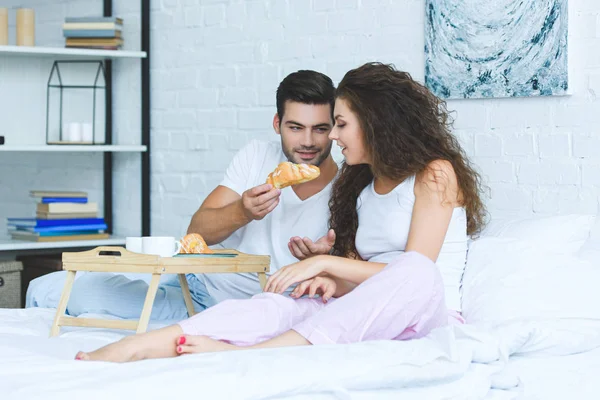 英俊的年轻人喂养美丽的微笑的女朋友与牛角面包在卧室里 — 免费的图库照片