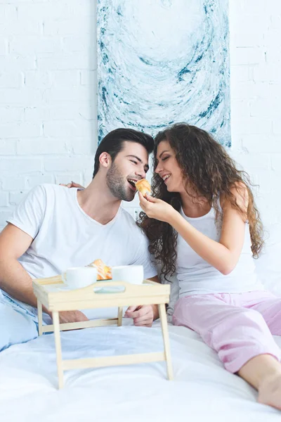 Hermosa Feliz Joven Mujer Alimentación Sonriente Novio Con Croissant Cama — Foto de stock gratis