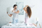 side view of happy young couple in pajamas fighting with pillows on bed  