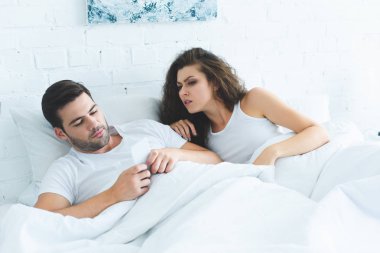 emotional young woman looking at boyfriend using smartphone in bed, relationship difficulties concept clipart