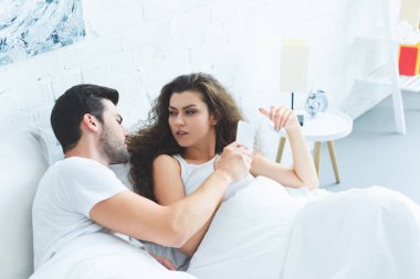 young couple holding smartphone and quarreling in bed, relationship difficulties concept clipart