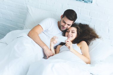 Genç çift smartphone birlikte yatakta kullanarak gülümseyen, yüksek açılı görünüş 