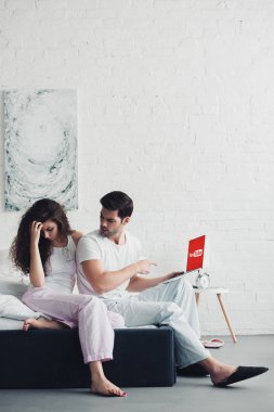 youtube Web sitesinde ekranı olan dizüstü bilgisayar, parmakla işaret eden adam yatakta oturan genç kadın 