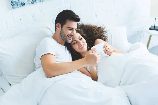 Mutlu Genç Çiftin Smartphone Birlikte Yatakta Kullanarak Yüksek Açılı Görünüş — Stok fotoğraf
