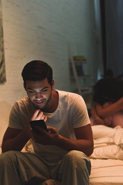Young Man Using Smartphone While Upset Girlfriend Sitting Bed Relationship — Free Stock Photo
