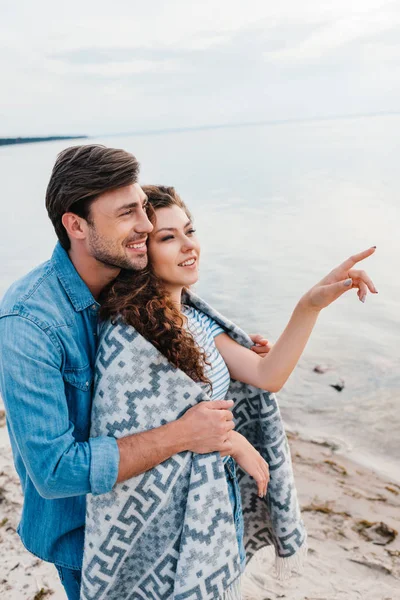 Happy Man Hugging Girlfriend Wrapped Blanket While She Pointing Sea — Free Stock Photo