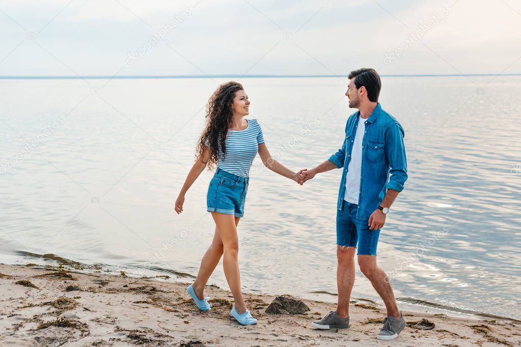happy couple holding hands and walking on beach near sea 