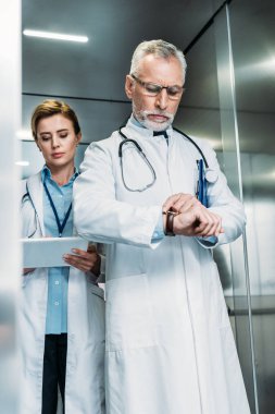 kendine güvenen orta yaşlı erkek doktor kadın meslektaşı dijital tablet arkasında hastane asansörde kullanma süre kol saati kontrol 
