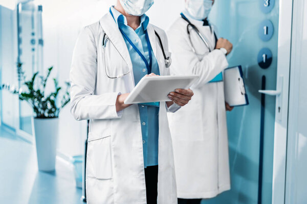 partial view of female doctor in medical mask using digital tablet while her male colleague standing behind with clipboard in hospital corridor 
