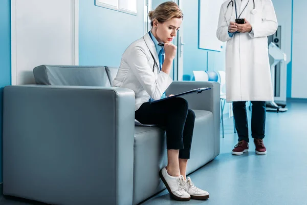 集中成年女医生坐与剪贴板在沙发上 而她的男性同事使用智能手机背后的医院走廊 — 图库照片