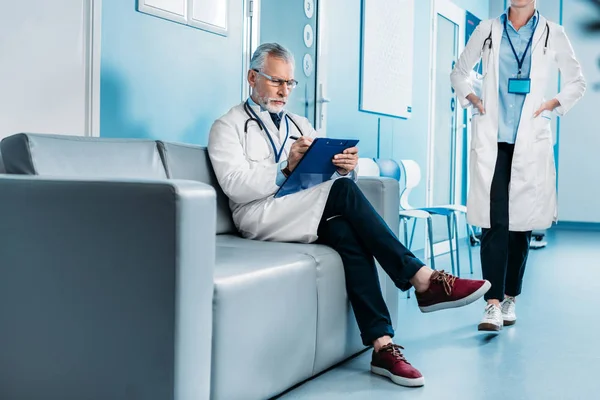 沉思中年男性医生写在剪贴板和坐在沙发上 而他的女同事走在医院走廊后面 — 图库照片