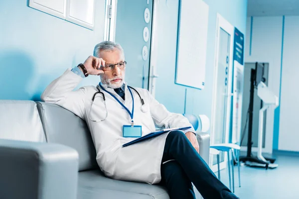 Tankeväckande Mellersta Åldern Manliga Läkare Med Urklipp Sittande Soffan Sjukhuskorridor — Stockfoto