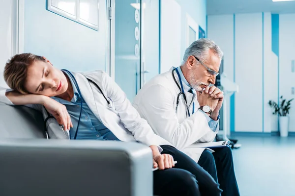 female doctor sleeping on couch while her male colleague sitting behind in hospital corridor