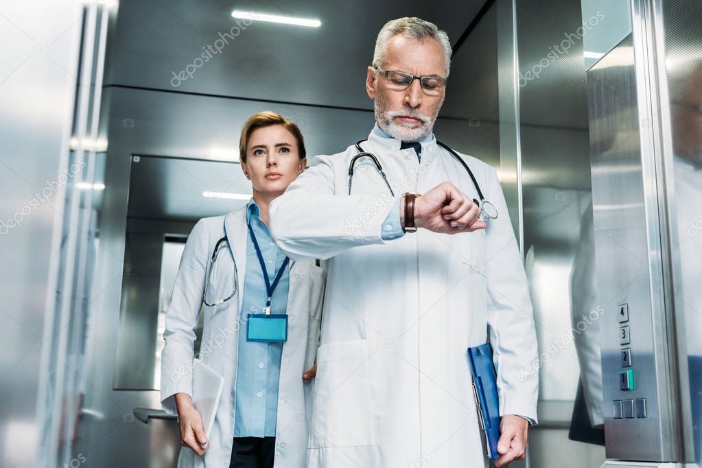 serious middle aged male doctor checking wristwatch while his female colleague standing behind in hospital elevator 