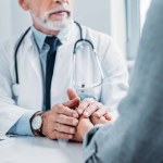 Vista parcial del médico masculino de mediana edad animando y tomándose de la mano de la paciente femenina en la mesa en el consultorio