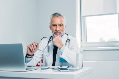 Gözlük tutan ve ofiste dizüstü bilgisayar ile tablo kameraya bakarak mutlu olgun erkek doktor 