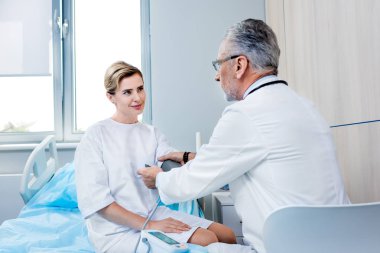 Orta yaşlı erkek doktor hastane odasında kadın hasta el basınç ölçme aygıtı koyarak