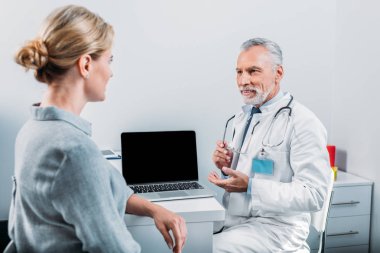 boş ekran ile laptop yakınındaki ofisinde oturan Bayan hasta için işaret olgun erkek doktor gülümseyen 