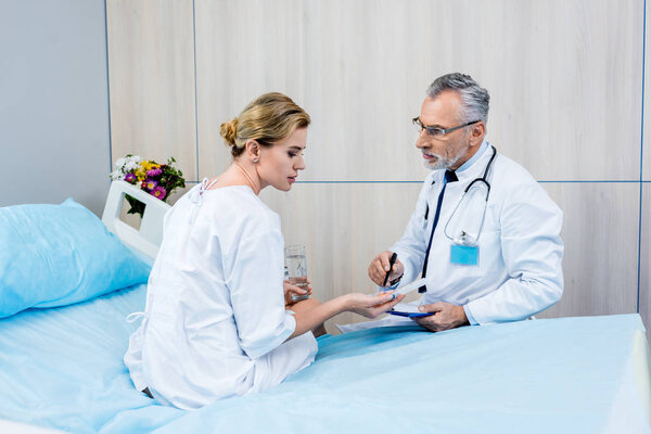 serious middle aged male doctor with stethoscope over neck pointing at pills to female patient in hospital room 