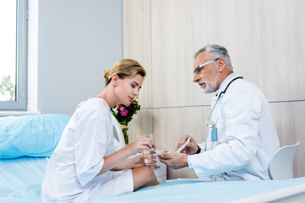 adult female patient with glass of water taking pills from mature male doctor in hospital room