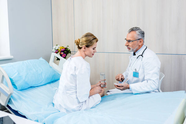 middle aged male doctor with stethoscope over neck pointing at pills to female patient in hospital room 