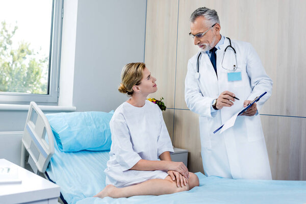 male doctor with stethoscope over neck pointing at clipboard to female patient in hospital room 